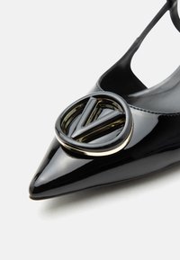 Μαύρο παπούτσι από πατεντ δέρμα με μυτερή μύτη, φλατ, με μεγάλο μαύρο και χρυσό κυκλικό διακοσμητικό έμβλημα στη μύτη.