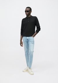 Polo preta de manga comprida, jeans slim azul claro e sapatilhas bege; o modelo usa óculos de sol pretos e está em frente a um fundo neutro.