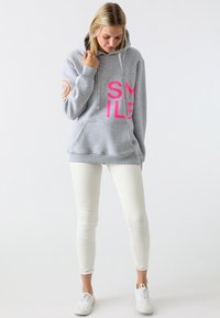 Felpa grigia con un grande grafico rosa "SMILE", cappuccio con cordini, tasca frontale e polsini a costine, abbinata a jeans skinny bianchi e sneakers bianche.