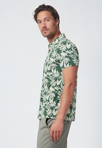 Camicia verde a maniche corte con motivo a foglie bianche, design con colletto e orlo dritto. Indossata con pantaloni grigi che presentano un logo discreto.
