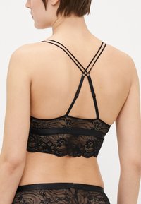 Svart blonderbralette med bølget blondekant, delikate blomsterdesign og justerbar bakdesign med doble stropper for støtte.