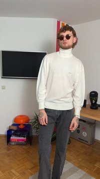 Cream turtleneck sweater with ribbed cuffs and embroidered logo, paired with dark grey flared jeans, displayed in a room with decor accents.