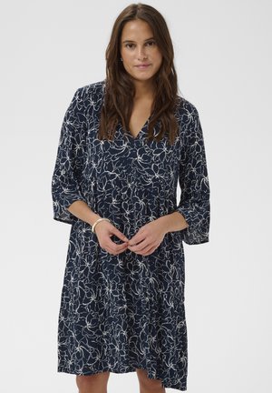 Ženska v mornarsko modri obleki do kolen z belim cvetličnim vzorcem, V-izrezom in tremi četrtinastimi rokavi, stoji z dvema prepletenima rokama.