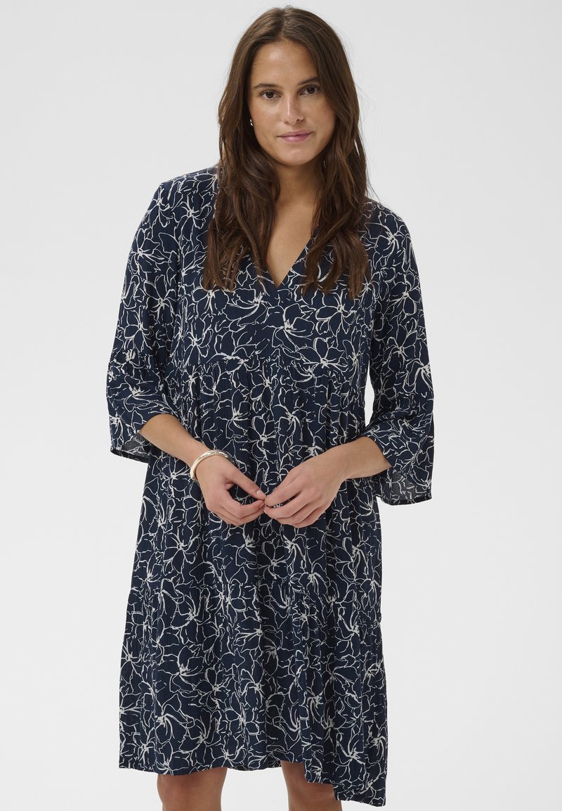 Sieviete, tērpusies ceļgalu garuma tumši zilā kleitā ar baltu ziedu kontūru rakstu, V veida kakla izgriezumu un trīs ceturtdaļu piedurknēm, stāv ar saliktām rokām.