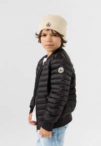Giacca a vento nera con polsini a costine, chiusura con zip e patch del logo sulla manica. Abbinata a un berretto in maglia color crema e una camicia bianca sotto.