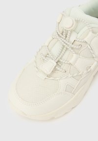 Tejszín színű sneaker hálós és bőr panelekből, gumis, nem kötős fűzőkkel, amelyek kerek zárókapoccsal rendelkeznek, fehér háttéren.