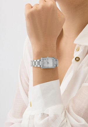Silver rectangular wristwatch with crystal details worn on left wrist over white long-sleeve shirt with gold buttons.