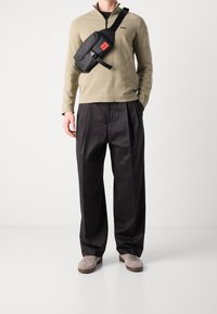 Man som bär en beige zip-up-tröja, svarta vida byxor, beige sneakers och en svart crossbody-väska med röd etikett.