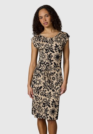 Frau mit lockigem Haar, die ein beiges knielanges Kleid mit schwarzen abstrakten Blumenmustern und einem Taillengürtel trägt, steht vor einem einfarbigen Hintergrund.