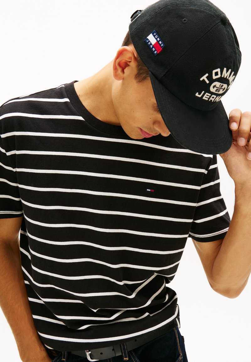 Homme portant un T-shirt rayé noir et blanc et une casquette noire Tommy Jeans, regardant vers le bas et ajustant la casquette d'une main.