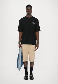 T-shirt en coton noir à manches courtes avec imprimé blanc, shorts beiges avec poches, chaussettes noires et mocassins noirs. Tenant un denim bleu.