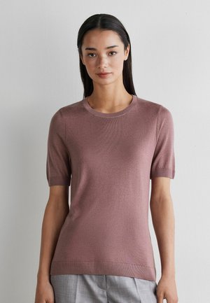 Vrouw met steil donker haar, gekleed in een mauve gebreide top met korte mouwen en grijze broek, staand tegen een effen lichte muur.