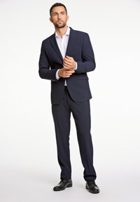 Abito blu navy con blazer aderente e pantaloni con fronte piatto, abbinato a una camicia bianca. Scarpe eleganti completano l'insieme.