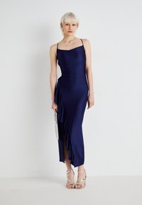 Navy satijnen jurk met dunne spaghettibandjes, met een gerimpeld detail aan de zijkant en een zijsplit. In combinatie met zilveren strappy hakken en een clutch.