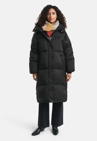 Vrouw met krullend haar, gekleed in een lange zwarte pufferjas, een meerkleurige sjaal, zwarte wijde broek en zwarte enkellaarzen, staande tegen een witte achtergrond.