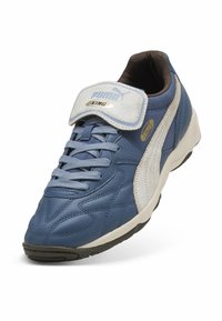 Sapatilha atlética Puma King em azul e branco com atacadores e uma tira de Velcro, exibindo detalhes de costura e sola texturizada.