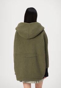 Sudadera oversized verde oliva hecha de felpa texturizada, con una gran capucha y puños de canalé negros. La vista trasera muestra un diseño minimalista.