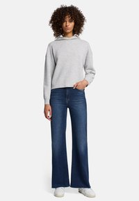 Felpa cropped grigia chiara con cappuccio abbinata a jeans a gamba larga blu scuro e sneakers bianche, che mostrano una combinazione di outfit informale.