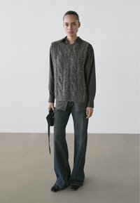 Brown knitted sleeveless sweater over a dark shirt, paired with flared blue jeans and black shoes. Textured and layered appearance.