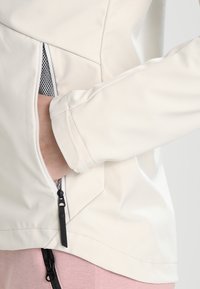 Main d'une personne dans la poche latérale zippée d'une veste blanc cassé, montrant la tirette noire du zip et un pantalon rose clair avec détail de zip noir.