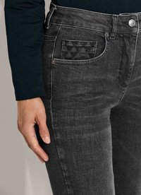 Close-up van een hand nabij de voorzak van vervaagde zwarte jeans, met een klein zwart patroonlabel in de zak.