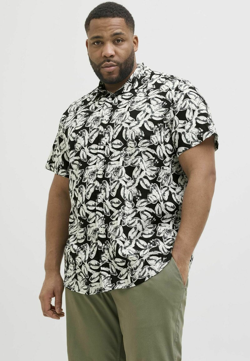 Schwarzes Kurzarm-Hemd mit weißen Blattmustern; mit Kragen und lässiger Passform. Kombiniert mit olivgrünen Hosen.