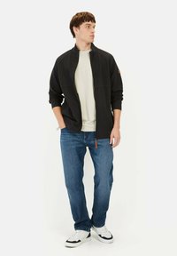 Zwarte zip-up jack met een hoge kraag en ribgebreide manchetten, gedragen over een lichtbeige t-shirt, gecombineerd met blauwe jeans en witte sneakers.