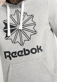 Šedá bavlněná mikina s kapucí, která má velký černý design ve tvaru hvězdného výbuchu a logo "Reebok" tištěné výraznou černou barvou. Obsahuje bílé šňůrky a přední kapsu.