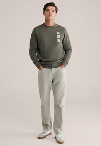 Donkergroen sweatshirt met een voorzak, ronde hals en witte grafische details, gecombineerd met lichtgrijze jeans en neutraal gekleurde sneakers.