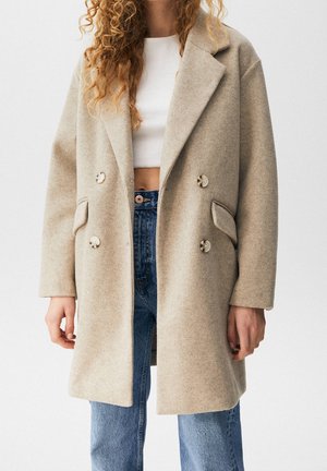 Kobieta w beżowym, wełnianym, dwurzędowym płaszczu, na białym topie z odsłoniętym brzuchem i niebieskich dżinsach, stoi na tle jednolitego białego tła.