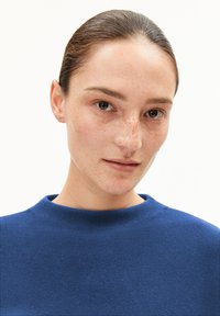 Blauer Strickpullover mit hohem Rundhalsausschnitt. Der Stoff hat eine strukturierte Oberfläche. Das Model hat einen neutralen Gesichtsausdruck und sichtbare Sommersprossen auf der Haut.