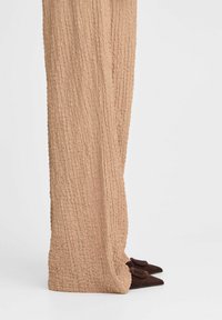 Texturierte, beigefarbene weit geschnittene Hosen mit einem zerknitterten Design. Kombiniert mit dunkelbraunen, spitzen Schuhen mit Schleifenakzenten.