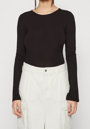 Vrouw draagt een getailleerd zwart shirt met lange mouwen, ingestopt in een witte hoge taille rok met voorzakken en riemlussen.