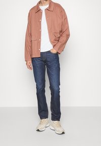 Muž v hnědo-oranžové bundě, bílem tričku, tmavě modrých džínách a béžových teniskách stojí na jednofonním bílém pozadí.