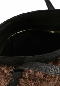 Bolso de mano de cuero negro con exterior de textil marrón rizado. Interior negro con un detalle de cremallera. Forma redondeada y asas cortas.