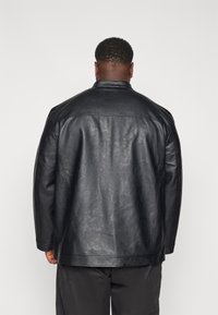 Chaqueta de cuero negra con un acabado texturizado, que presenta un cuello alto y mangas estructuradas. Diseño simple sin herrajes o patrones visibles.