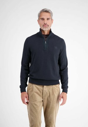 Man with gray hair and beard wearing a black quarter-zip pullover and light brown corduroy pants standing against a plain white background.