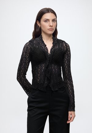 Femme aux cheveux raides et foncés portant un chemisier en dentelle noire transparente et un pantalon noir, debout devant un fond clair uni.