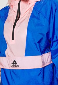 Blå og pink vindjakke med kvart-zip krave, elastiske håndled og et Adidas-logo på forsiden. Har en glat, let tekstur.