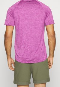 Zadní pohled na osobu v krátkém rukávu fialového heatherového sportovního trička a olivově zelených šortkách.