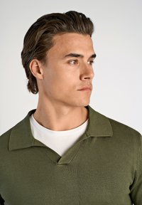 Olivgrüner Pullover mit V-Ausschnitt und kurzem Kragen, aus strukturiertem Strickmaterial, über einem weißen Rundhals-T-Shirt getragen.