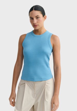 Femme aux cheveux foncés portant un haut sans manches bleu côtelé et un pantalon plissé beige, debout devant un fond clair uni.