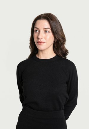 Schwarzer, langärmeliger Pullover aus weichem Material mit rundem Halsausschnitt und taillierter Silhouette; Modell steht mit den Händen hinter dem Rücken vor einem neutralen Hintergrund.