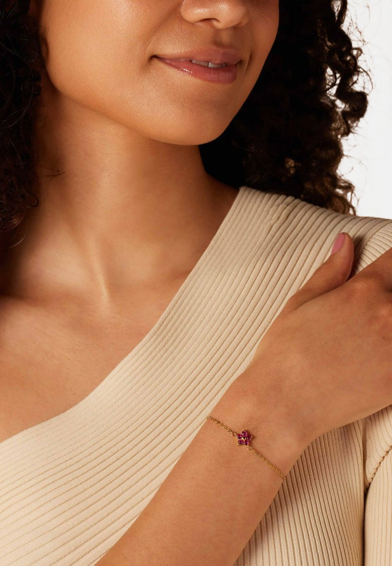 Gouden kettingarmband met een flower design en roze edelstenen, gedragen om een licht gebruinde pols tegen een textuurachtige beige stof achtergrond.