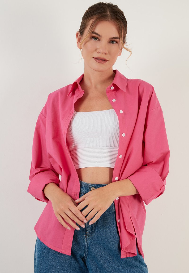 Pink button-up shirt made of cotton, featuring a relaxed fit, long sleeves, and white buttons, paired with a white cropped top and blue jeans.