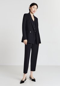 Blazer nero su misura con due bottoni, abbinato a un gilet coordinato e pantaloni cropped. Tacchi slingback neri completano l'outfit.