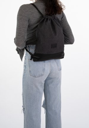 Person, die hellblaue Jeans und ein graues Langarmshirt trägt, mit einem schwarzen Rucksack mit Kordelzug, von hinten gezeigt.