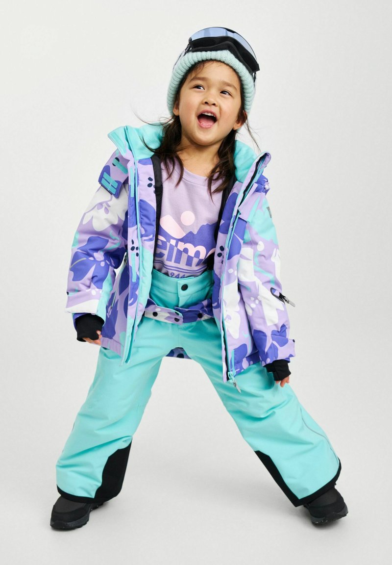 Tenue de ski colorée comprenant une veste violette à motifs, un pantalon aqua et un bonnet bleu clair. Inclut des lunettes de ski et des bottes noires.
