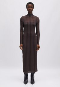 Durchsichtiges, langärmeliges, schwarzes Kleid mit vertikalen braunen Streifen, mit hohem Kragen und figurbetontem Schnitt, kombiniert mit schwarzen Knöchelstiefeln.