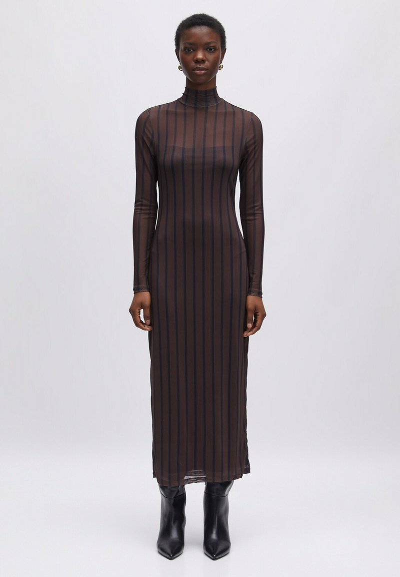 Durchsichtiges, langärmeliges, schwarzes Kleid mit vertikalen braunen Streifen, mit hohem Kragen und figurbetontem Schnitt, kombiniert mit schwarzen Knöchelstiefeln.